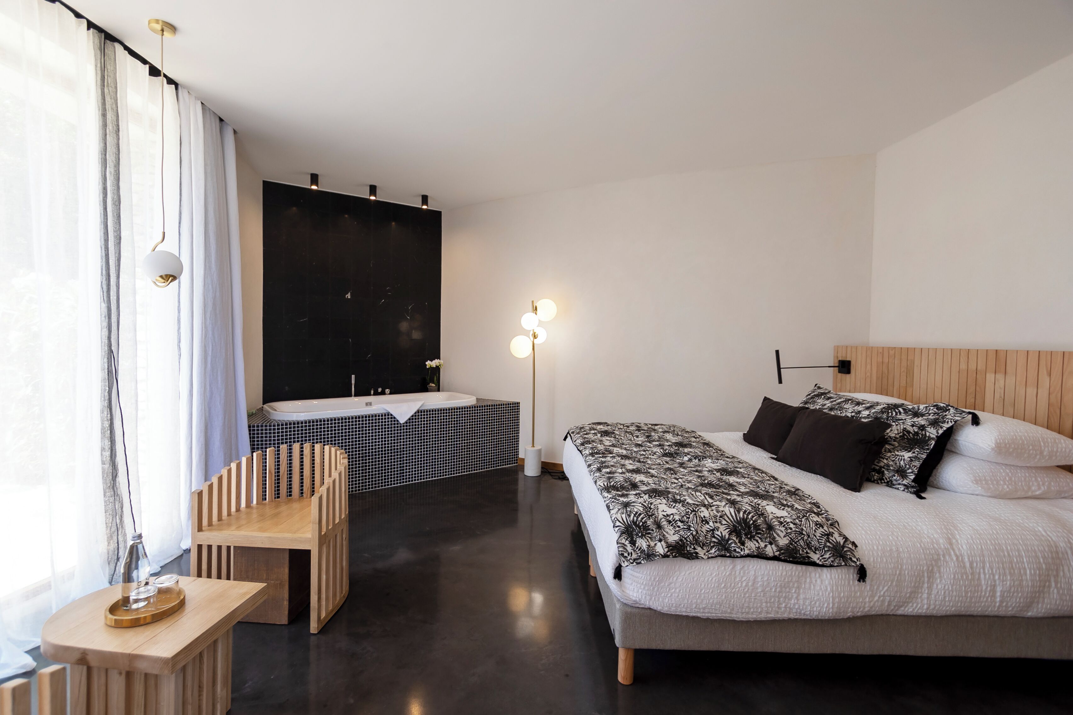 A modern bedroom with a large bed, a wooden side table, a small desk, a large window with curtains, a black wall, and a floor lamp.