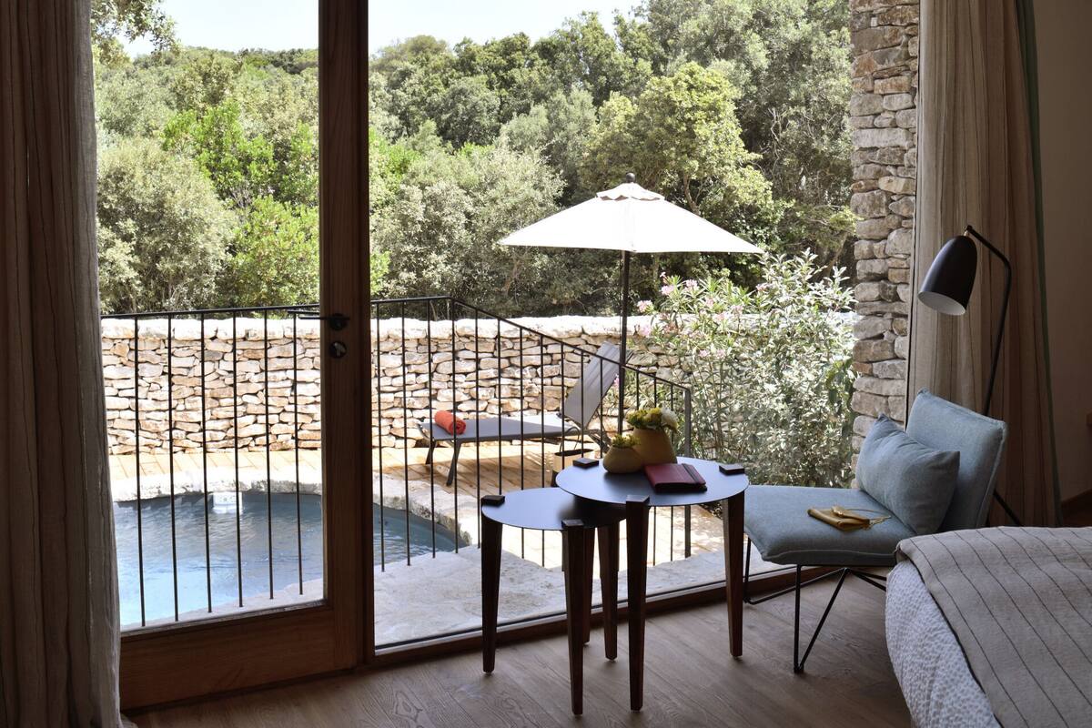 A balcony with a table, chairs, and a small pool, overlooking a stone wall and greenery.