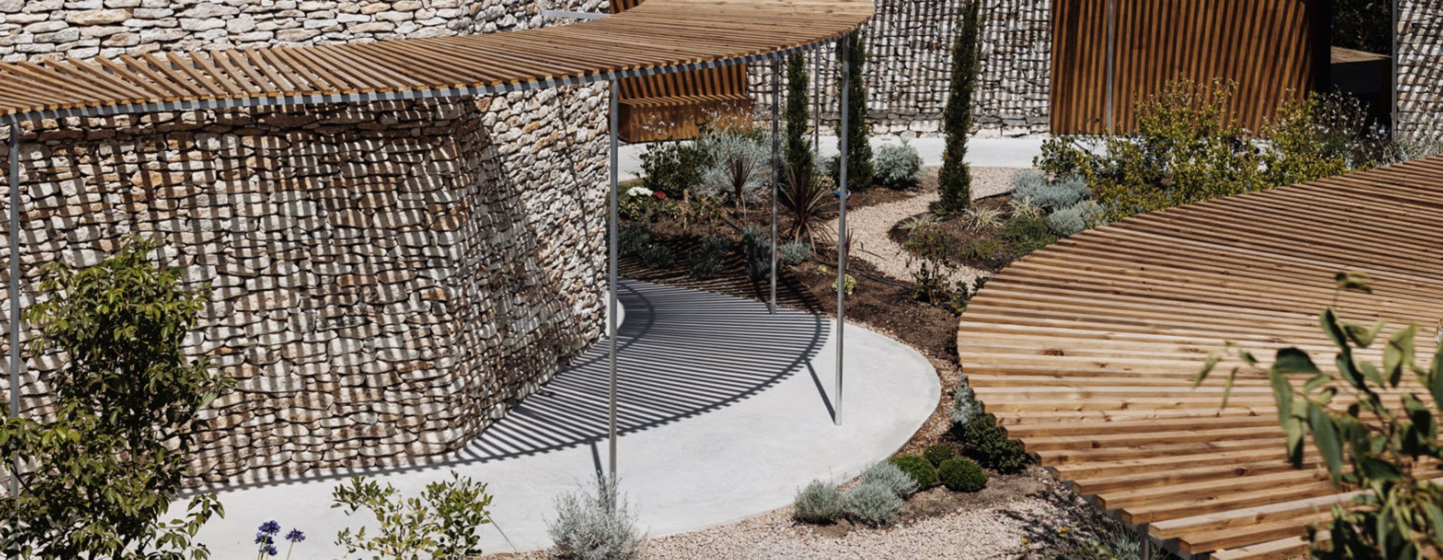 Un espacio exterior moderno con un muro de piedra, pérgolas de madera y un sendero curvo.