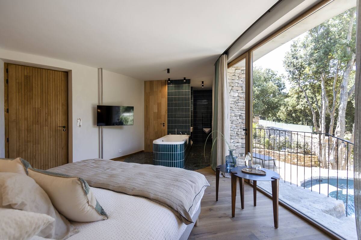 A modern bedroom with a large bed, a TV, a bathroom with a bathtub, and a balcony with a pool.