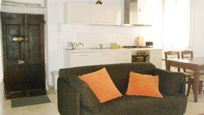 A living room with a dark sofa, two orange cushions, a wooden dining table, and a kitchen in the background.
