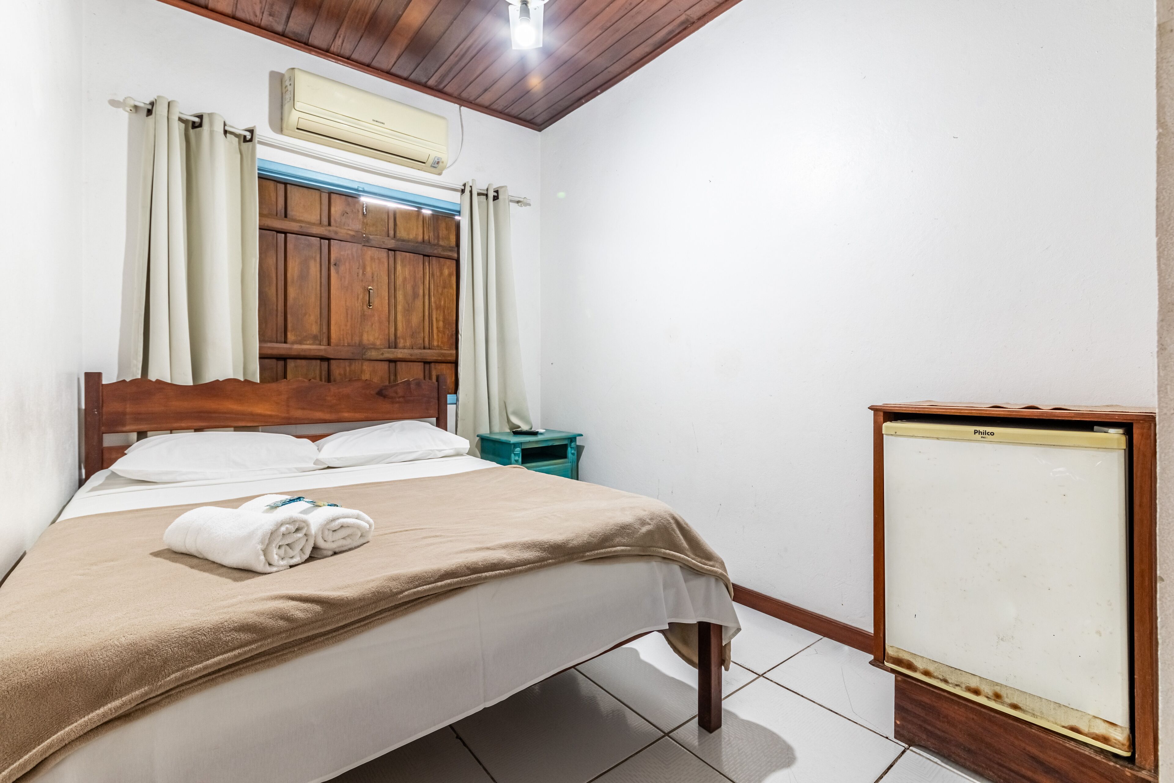 Una cama bien hecha con una manta color beige y toallas enrolladas, un cabecero de madera, una mesita de noche azul verdoso y un aire acondicionado blanco, además de un armario de madera con puerta blanca.