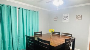 Un coin repas avec une table et des chaises en bois, des rideaux turquoise, un ventilateur de plafond et un panneau mural sur lequel on peut lire : « Rassemblez-vous autour de la table ».