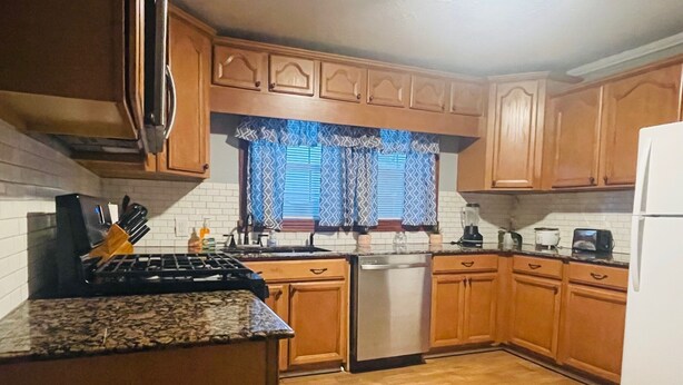Une cuisine avec des meubles en bois, un plan de travail en granit, une cuisinière, un lave-vaisselle et une fenêtre agrémentée de rideaux à motifs.