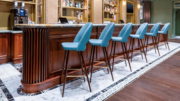 Un espace bar doté d’un sol en marbre, d’un comptoir en bois et de tabourets de bar bleus. On y trouve des étagères remplies de bouteilles et un téléviseur fixé au mur.
