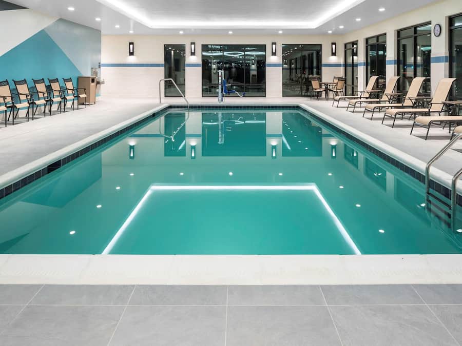 En stor indendørs swimmingpool med krystalklart vand, omgivet af liggestole og et reception område.