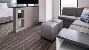 Une chambre d’hôtel moderne comprenant un lit, un téléviseur à écran plat, un canapé gris, un petit pouf et une table basse.