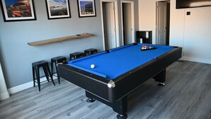 A pool table with a blue felt surface, black legs, and a white cue ball. There are two black stools and a wooden shelf above the table. - Spacious Riverhouse with private park/boat ramp (Parker)