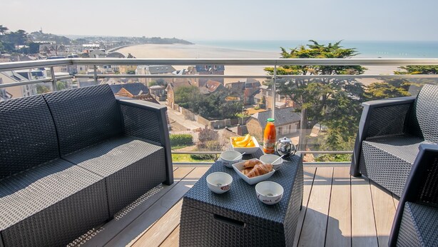 Een balkon met rieten meubels, een tafel met ontbijtspullen en uitzicht op strand en stadsbeeld.