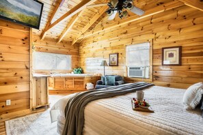 Ein gemütliches Blockhaus mit Holzvertäfelung, einem Bett mit Decke und einer Obstschale, einem Schreibtisch mit Lampe sowie einem Deckenventilator.