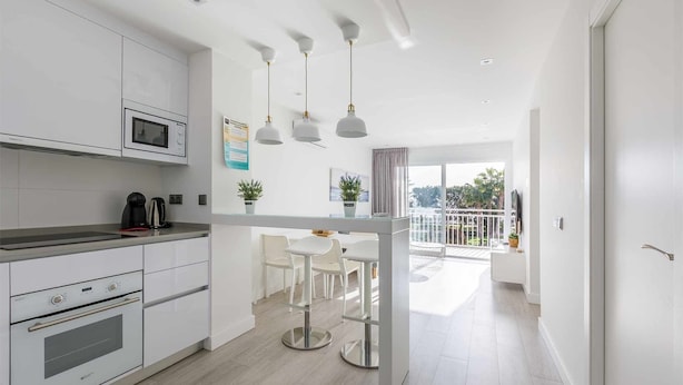 A modern kitchen with white cabinetry, a built-in microwave, and stainless steel appliances. There is a dining area with white chairs and a glass-top table. The room has a balcony with a view of the outdoors. - A&n Seafront Torre del Mar (Torre del Mar)