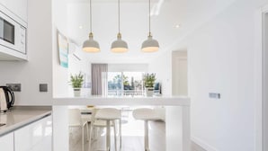 A modern kitchen with white cabinetry, a central island with bar stools, pendant lighting, and a view of the city through large windows. - A&n Seafront Torre del Mar (Torre del Mar)