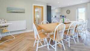 A dining area with a wooden table and white chairs, a kitchen in the background, and a view through an open door.