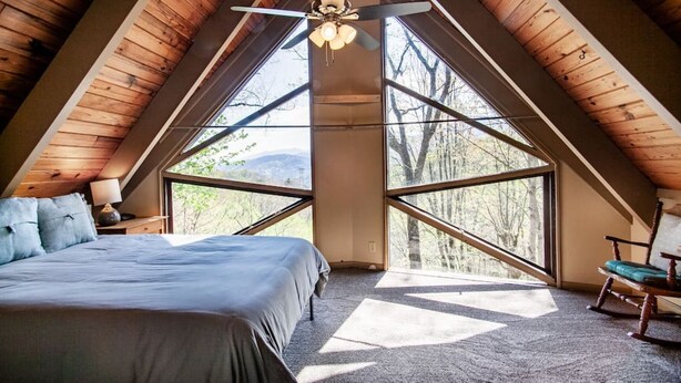 A bedroom with a sloped ceiling, a bed, a rocking chair, and large windows offering a view of trees and mountains.