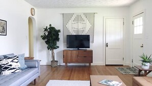 Una sala de estar con un sofá, una mesa de centro, un televisor sobre un mueble de madera, una planta en maceta y un tapiz con estampado.