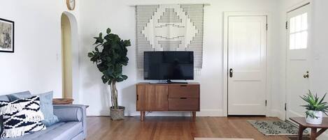 A living room with a sofa, a coffee table, a TV on a wooden cabinet, a potted plant, and a patterned wall hanging.