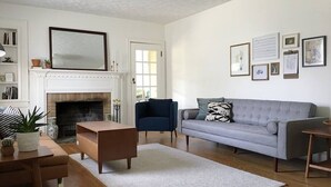 Una sala de estar con chimenea, un sofá gris, un sillón azul, una mesa de centro de madera y una alfombra blanca.