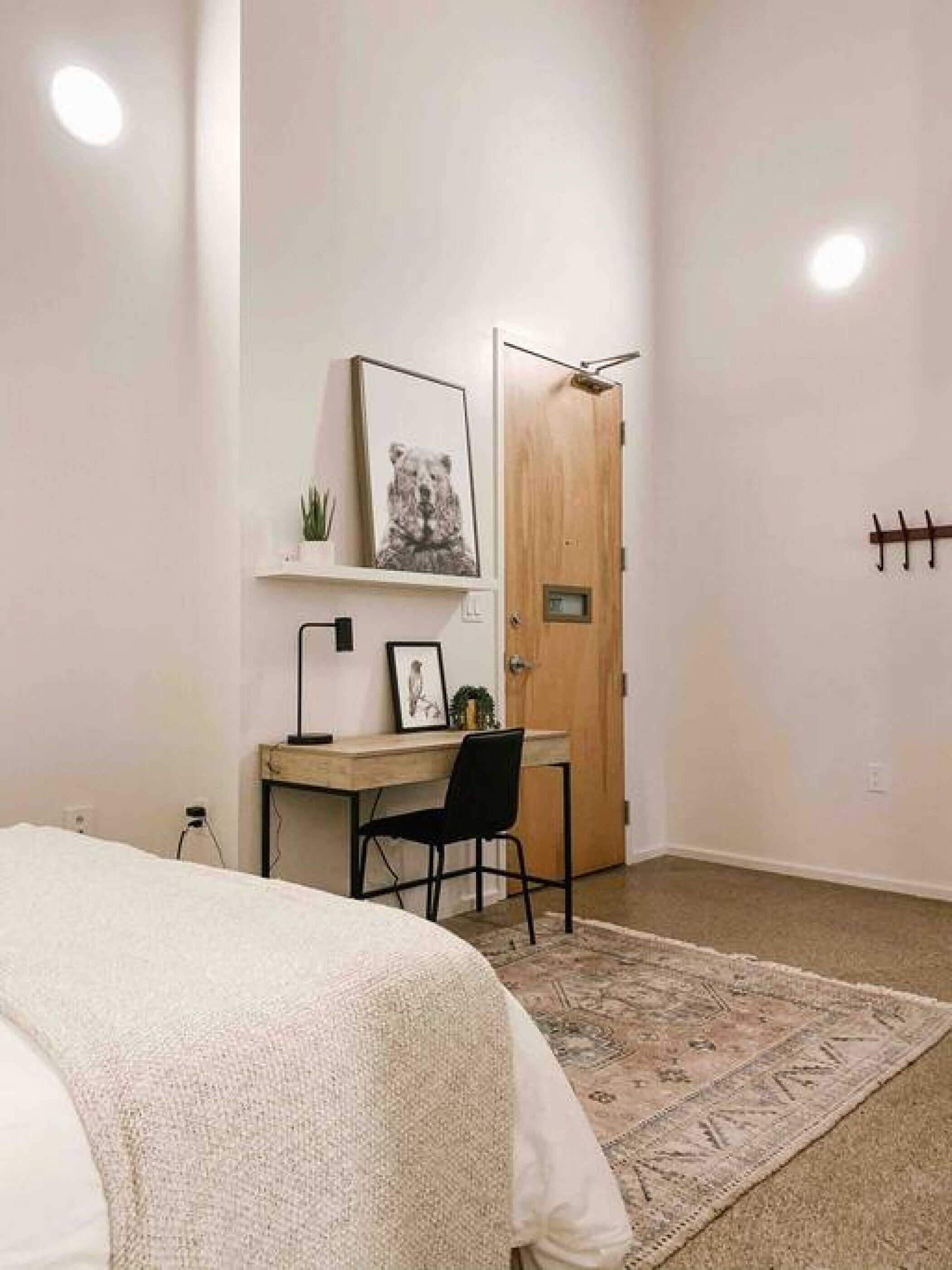 Un dormitorio con cama, escritorio, silla, puerta y perchero en la pared.