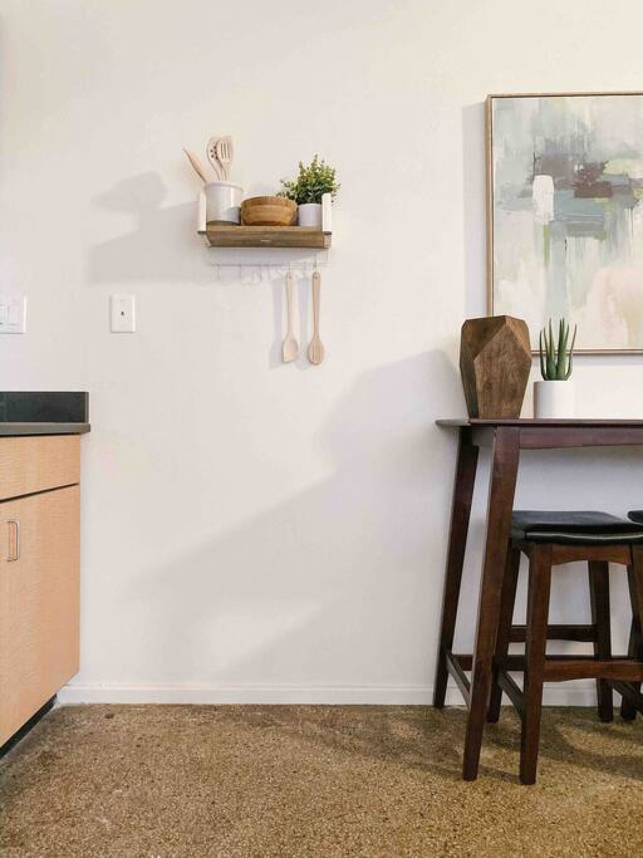 Una cocina con un armario de madera, una mesa de comedor con una silla, un estante con utensilios de cocina, y un cuadro enmarcado.