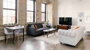 A spacious living room with large windows, a sofa, a white armchair, a coffee table, and a TV. - Modern Gay St Loft w/ View (Knoxville)