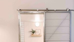 A room with a sliding door leading to a bathroom, a bed with a patterned headboard, and a small shelf with a plant and picture frame.