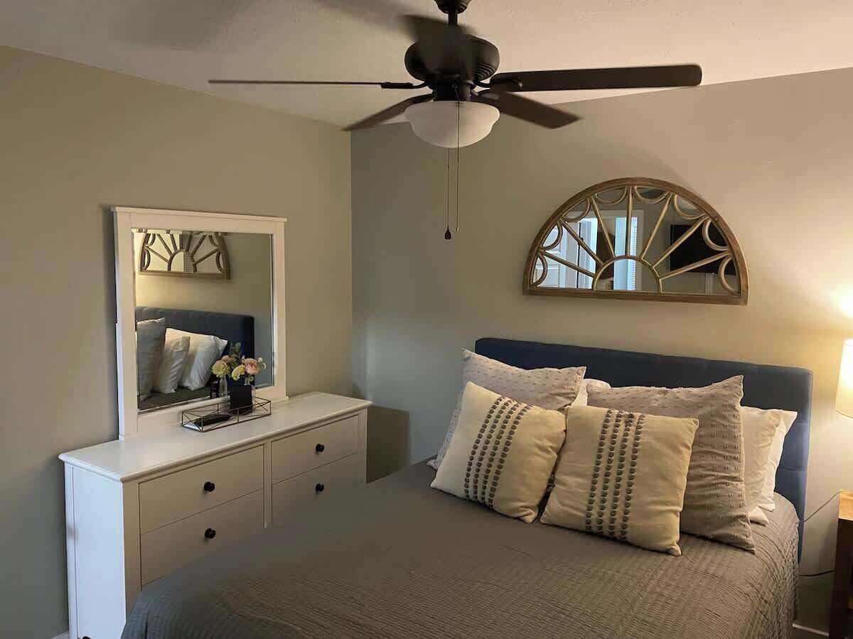 Een slaapkamer met een bed, een dressoir, een plafondventilator, een spiegel en een decoratief object aan de muur.
