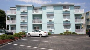A multi-story building with balconies, a parking lot, and a white car parked in front.