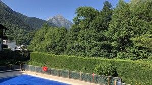 En swimmingpool med ett blått poolskydd, omgiven av ett trädäck och ett metallräcke, med en bakgrund av frodig grönska och berg.