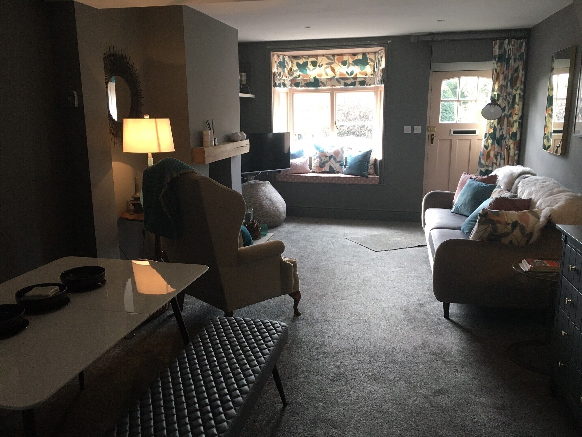 Een woonkamer met een bank, fauteuil en een eettafel. Er is een raam met gordijnen en uitzicht op de omgeving.