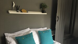 Cama bem arrumada com lençóis brancos e almofadas azul-turquesa, prateleira branca com objetos decorativos e porta que dá para outro quarto.