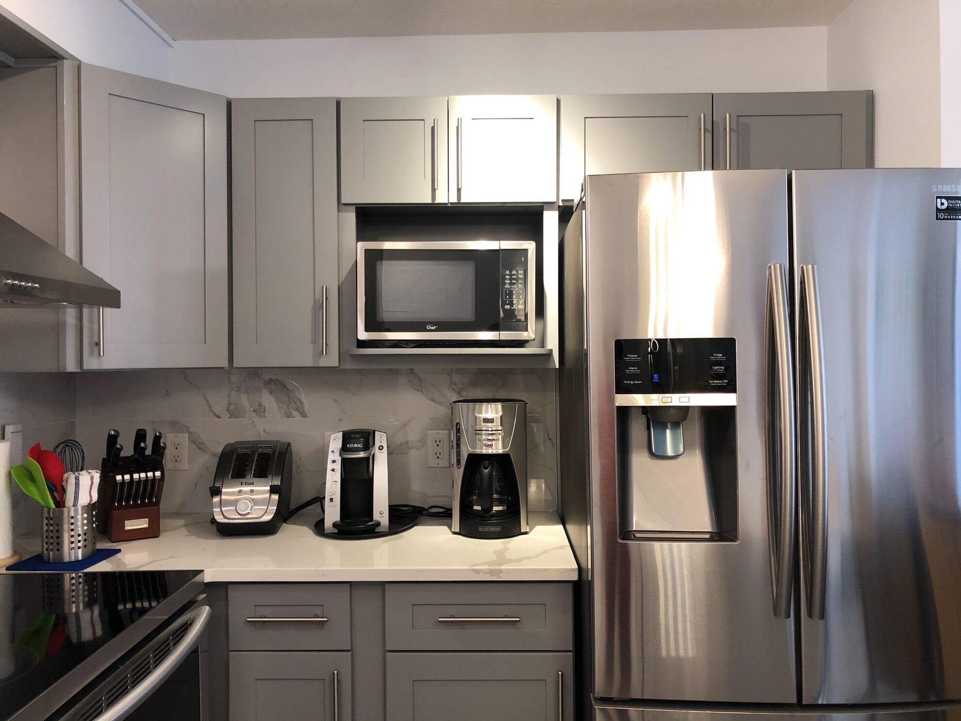 A modern kitchen with grey cabinets, a stainless steel refrigerator, a microwave, a coffee maker, and a toaster.