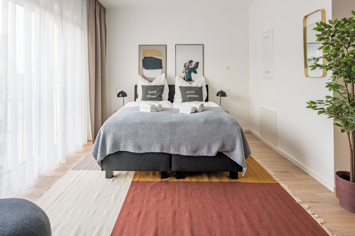 A modern bedroom with a bed, two pillows, a bedside table, a lamp, a plant, and a window with curtains.