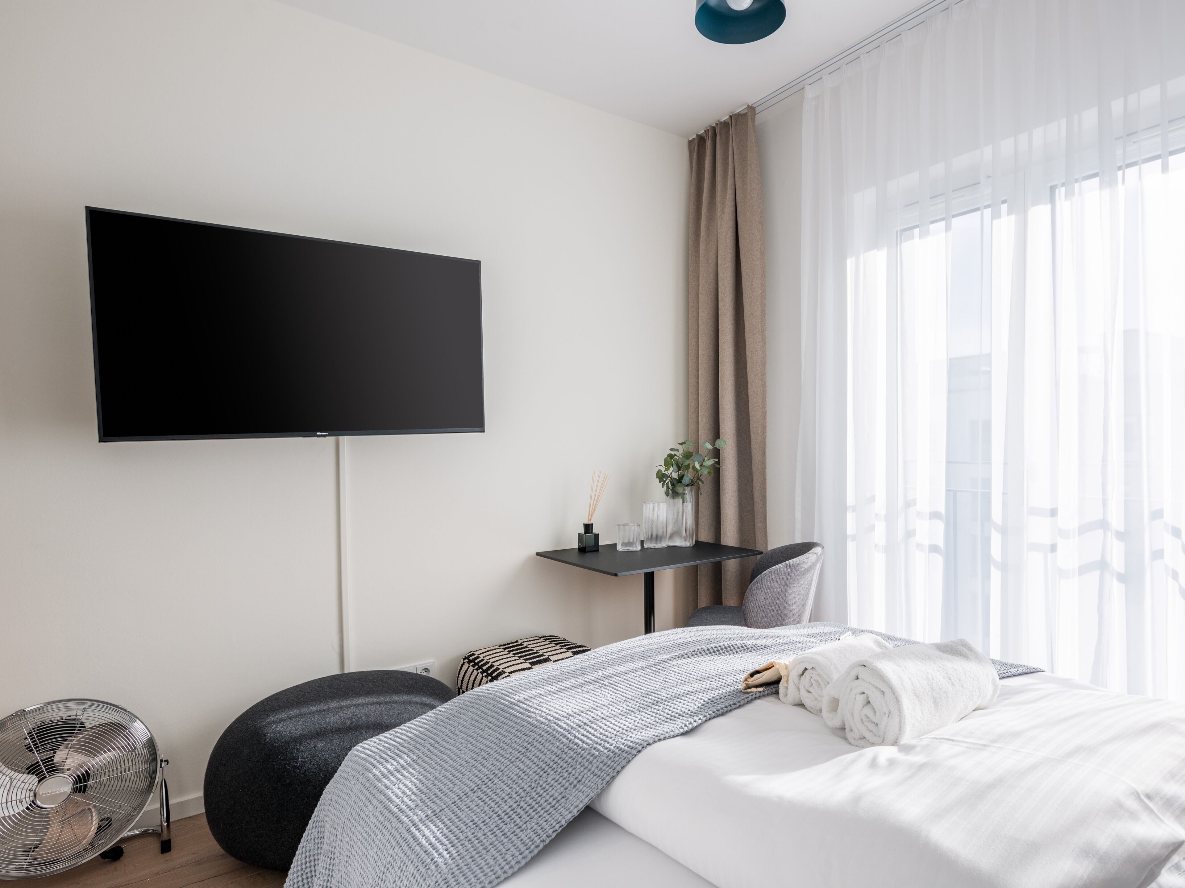 A modern hotel room with a flat-screen TV mounted on the wall, a small table with a plant, a fan, and a bed with a grey blanket.
