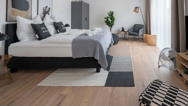 A modern bedroom with a bed, bedside tables, a wardrobe, a small side table, a chair, a fan, and a window with curtains. - Limehome Frankfurt Gutleutstraße (Frankfurt)