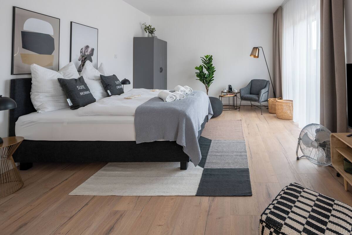 A modern bedroom with a bed, bedside tables, a wardrobe, a small side table, a chair, a fan, and a window with curtains.