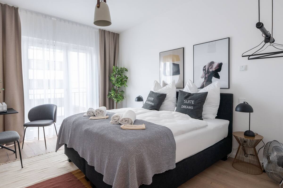 A modern hotel room with a large bed, a desk, a chair, and a plant.