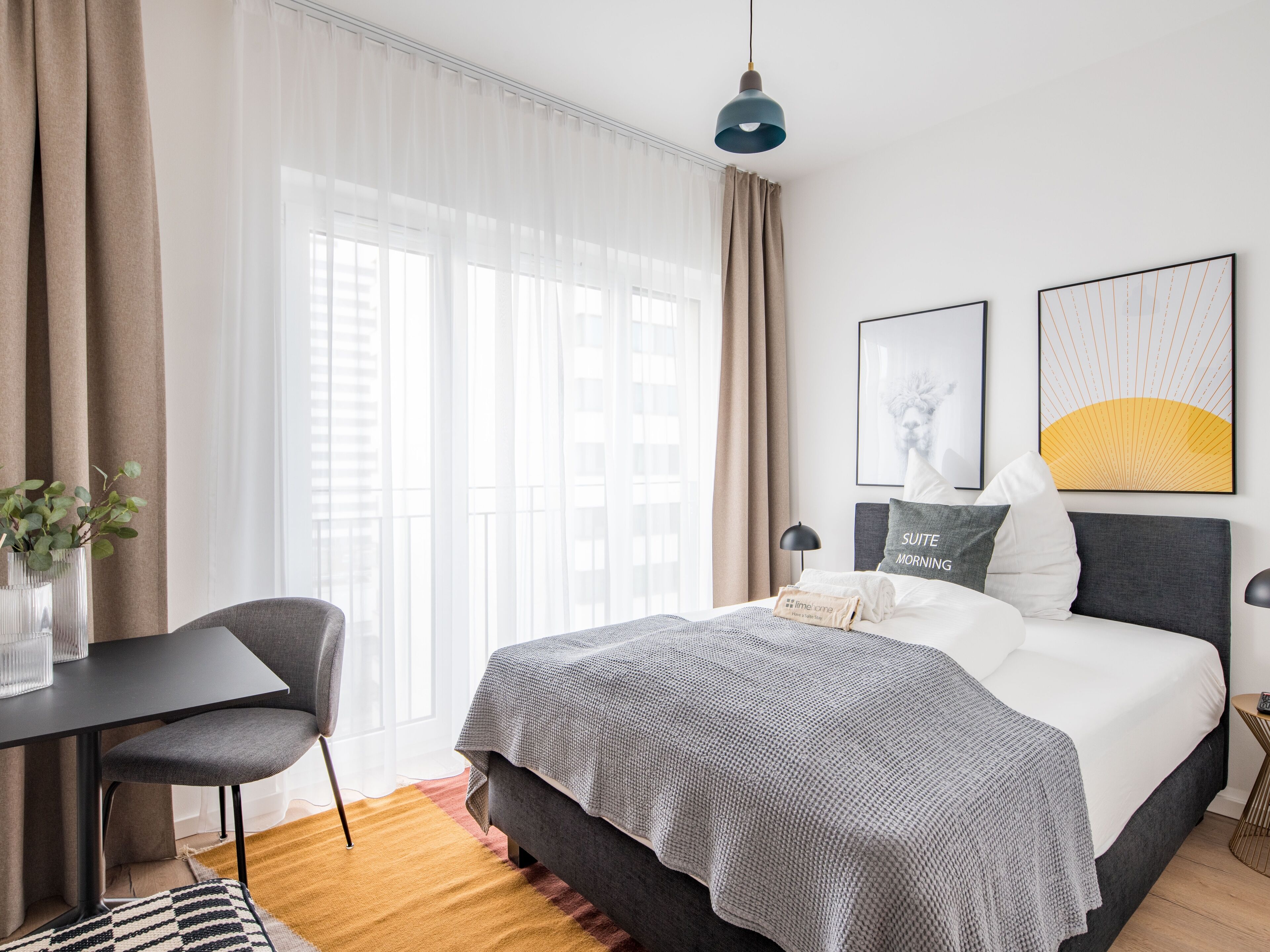 A hotel room with a bed, a desk, a chair, a lamp, and artwork on the wall.