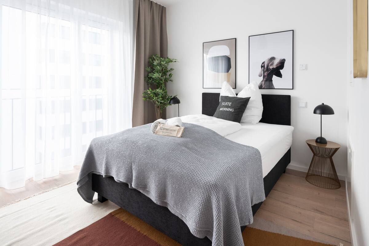 A modern bedroom with a bed, a bedside table, a lamp, and a window with curtains.