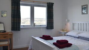 Ett sovrum med en säng, ett sängbord, en lampa, ett fönster med gardiner och utsikt över omgivningen.