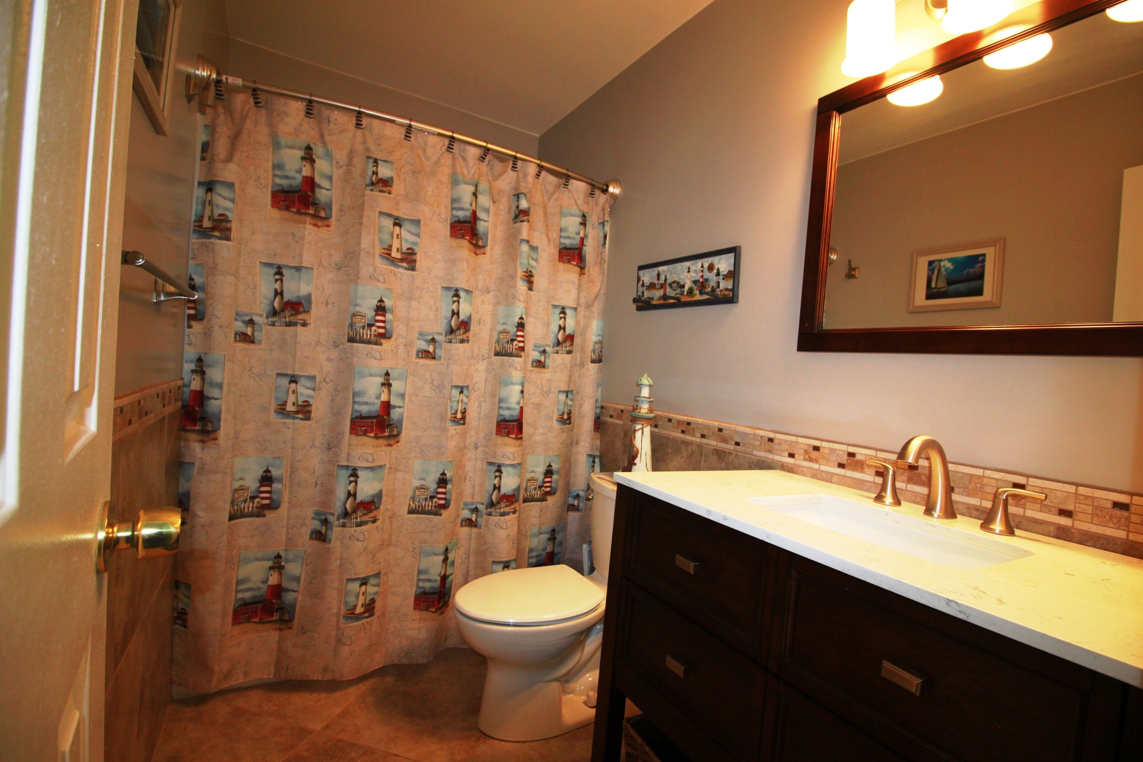 A bathroom with a shower curtain featuring a lighthouse pattern, a white toilet, a sink with a gold faucet, and a mirror above the sink.