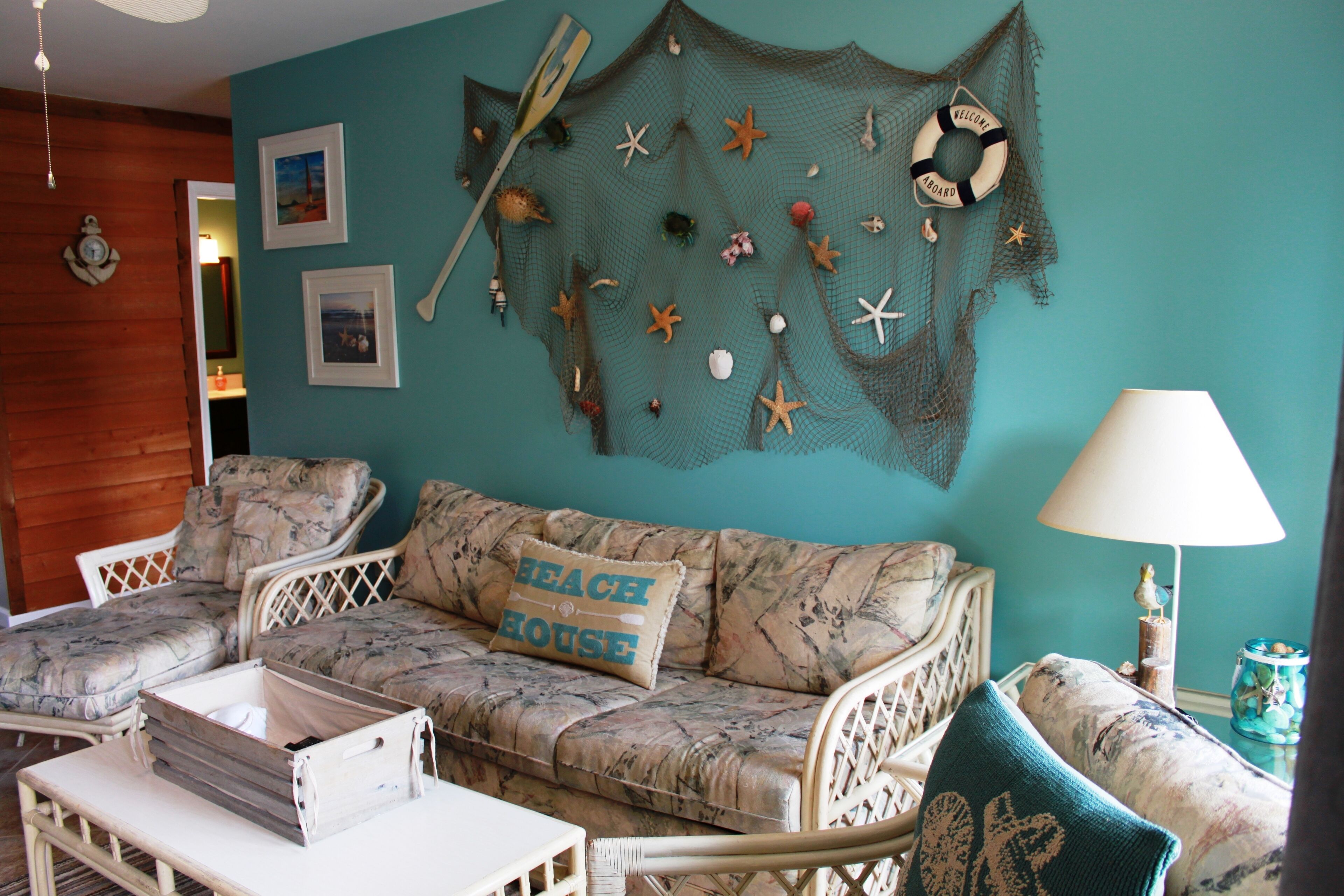 Une pièce avec un mur de couleur turquoise, une décoration murale tissée aux motifs marins, un canapé agrémenté d’un coussin orné de l’inscription « MAISON DE PLAGE », et une petite table avec une lampe.