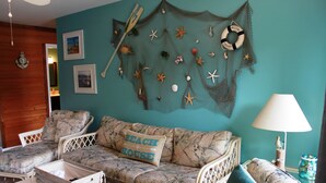 Ett rum med en turkosfärgad vägg, en vävd väggdekoration med snäckskal och sjöstjärnor, en soffa med en kudde med texten ”BEACH HOUSE”, en flätad stol och ett bord med en lampa.