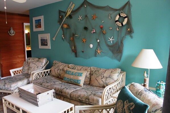 A room with a teal wall, a woven wall hanging with nautical elements, a sofa with a 'BEACH HOUSE' pillow, and a small table with a lamp.