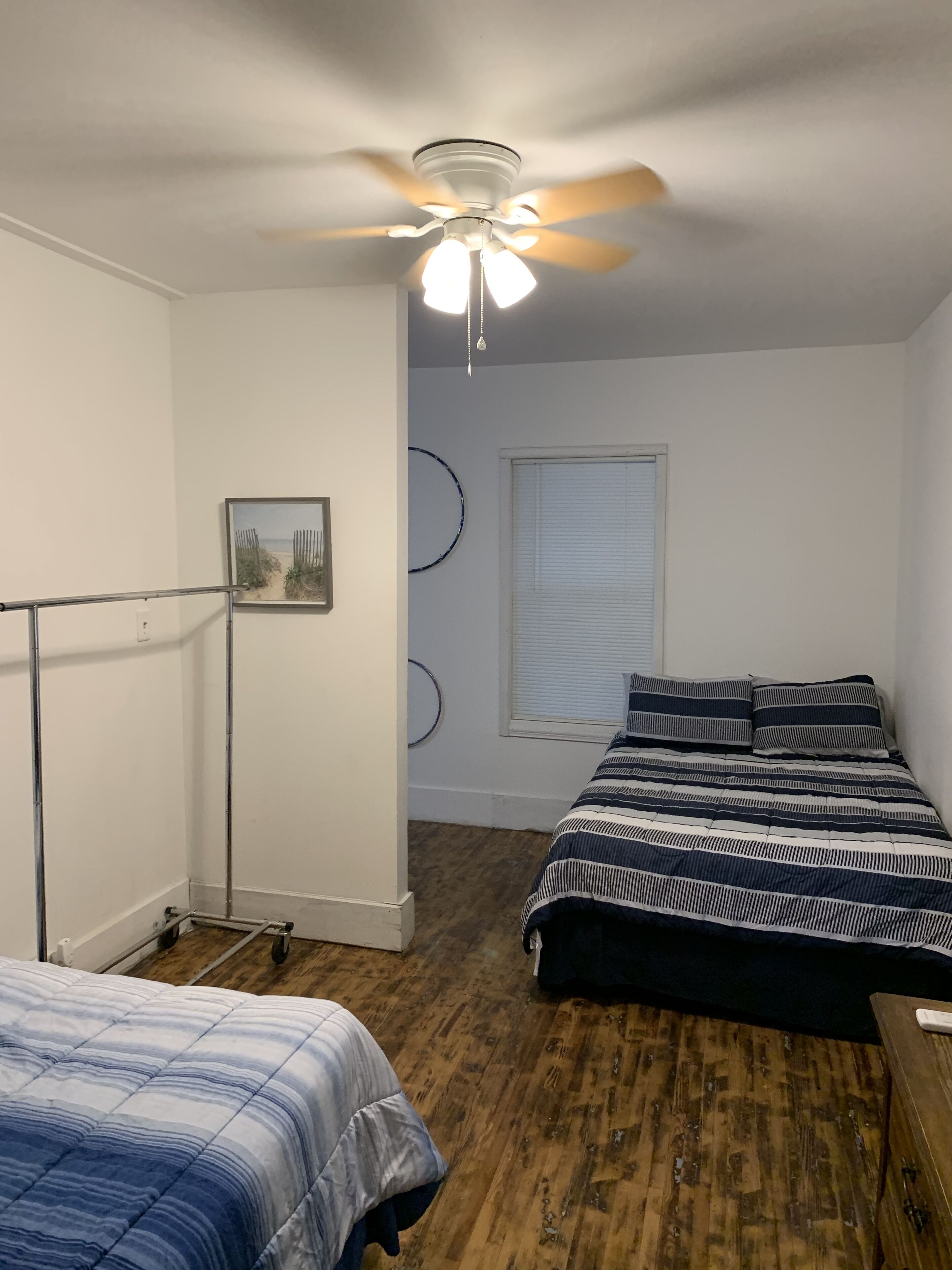 A bedroom with two beds, a ceiling fan, a window with blinds, and a wall-mounted circular decoration.