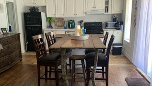 Zona pranzo con tavolo e sedie in legno, un frigorifero e pensili bianchi sullo sfondo.