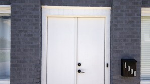 A white double door with a black handle and a keyhole, set in a brick wall with a mailbox displaying the number 1000.