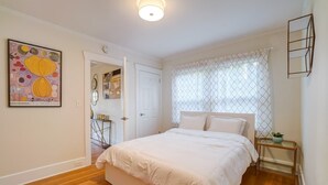 A bedroom with a large bed, a bedside table with a plant, a window with a patterned curtain, and a door leading to another room. - Fresh Modern Home in the Alberta Arts District! (Portland)