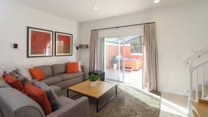 A modern living room with a grey sofa, orange cushions, a wooden coffee table, and a large sliding glass door leading to an outdoor area.