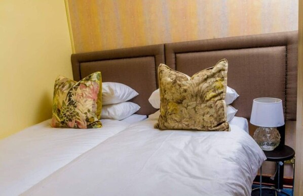 Une chambre d’hôtel avec un lit, deux coussins à motifs floraux, une lampe de chevet et un mur uni.