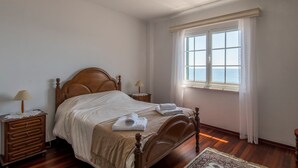 Ein Schlafzimmer mit einem hölzernen Bett, zwei Nachttischen mit Lampen, einem Fenster mit Meerblick und einem gemusterten Teppichboden.